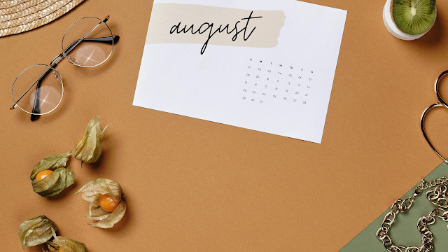 Vista superior de uma mesa com uma folha de calendário do mês de agosto, óculos redondos, frutas physalis, meio kiwi, tesoura dourada em forma de coração e uma corrente dourada, sobre fundo em tons de castanho e verde.