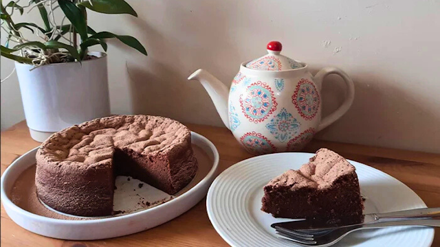 Bolo intenso de chocolate para o Dia Mundial do Cacau Bolo caseiro de chocolate rico em cacau, servido numa forma branca com uma fatia no prato, acompanhado por um bule de chá decorado e uma planta ao fundo, num ambiente acolhedor.