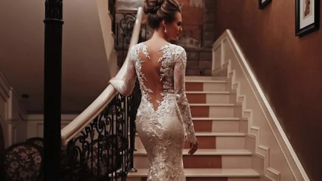 Noiva a subir escadas com vestido de noiva rendado, mangas compridas e costas abertas, num ambiente clássico e elegante