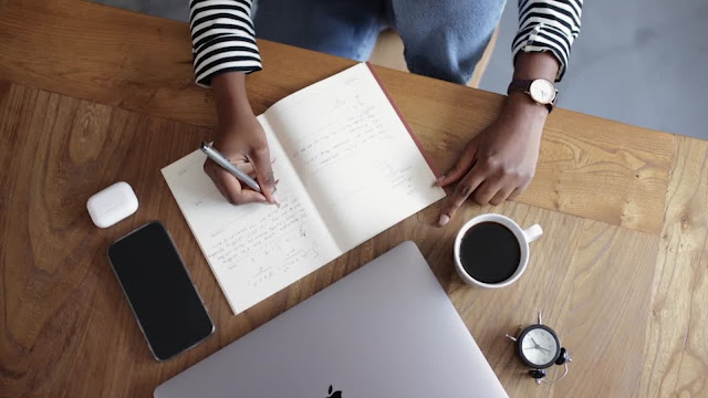 Pessoa a escrever num caderno num ambiente de trabalho, com portátil, café e smartphone, a planear uma análise SWOT pessoal.