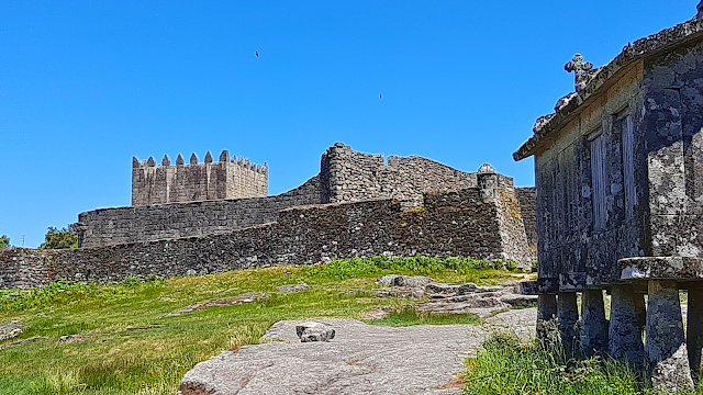 Vista do Castelo de Lindoso com as muralhas de pedra ao fundo e um espigueiro tradicional em primeiro plano num dia de céu azul.