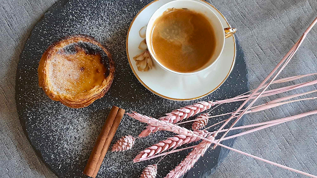 Chávena de café expresso acompanhada de um pastel de nata, polvilhado com açúcar em pó, sobre um prato de ardósia. Ao lado, um pau de canela e ramos secos cor-de-rosa decoram a composição.