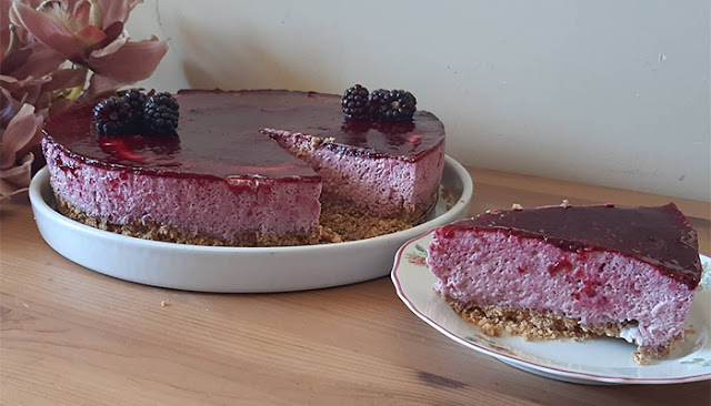 Tarte de amoras com base de bolacha e cobertura brilhante de polpa de amora, servida numa travessa branca com uma fatia à parte num prato floral.
