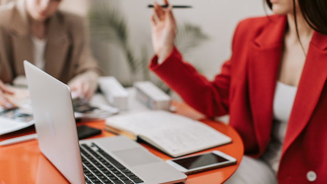 Duas mulheres em reunião profissional, uma a comunicar de forma assertiva enquanto aponta com uma caneta, com computador portátil, caderno e telemóvel sobre a mesa