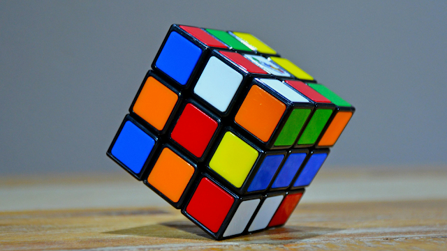 Um close de um Cubo Mágico 3x3 baralhado, apoiado num ângulo sobre uma mesa de madeira. O cubo tem faces coloridas com autocolantes de plástico (vermelho, azul, verde, amarelo, laranja, branco) dispostos aleatoriamente. O fundo é de uma cor cinzenta sólida.