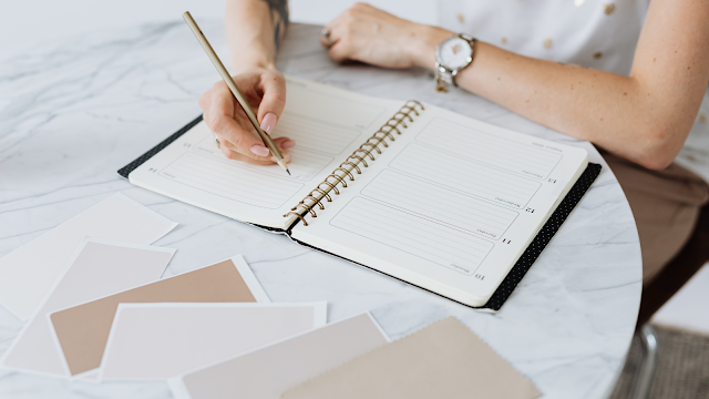 Planeamento e Organização Pessoal com Agenda Fotografia em plano aproximado de uma pessoa a escrever numa agenda aberta sobre uma mesa de mármore branco. A pessoa segura um lápis dourado e tem os braços apoiados na mesa, revelando um relógio de pulso prateado. Ao lado da agenda, encontram-se alguns cartões em tons pastéis e terra. O ambiente é iluminado, transmitindo uma sensação de foco e organização.