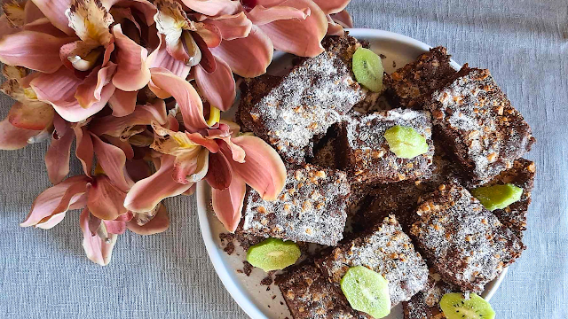 Prato com brownies de chocolate polvilhados com açúcar e avelã picada, decorado com pétalas verdes, ao lado de flores rosa sobre uma toalha cinzenta.