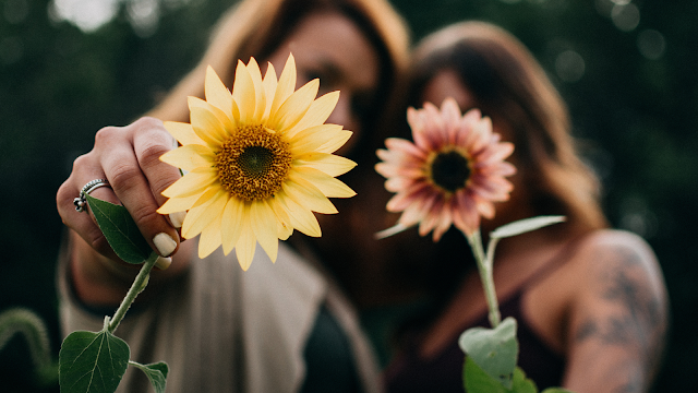 Duas mulheres seguram flores amarelas e rosadas, simbolizando a amizade e a ligação feminina, com foco nas flores e fundo desfocado.