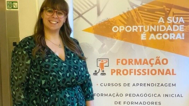 Mulher sorridente, de óculos, em contexto de formação profissional, em frente a um painel informativo sobre cursos de aprendizagem e formação pedagógica de formadores.