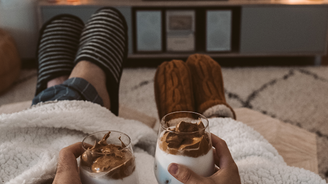 Perspetiva em primeira pessoa de duas pessoas sentadas num sofá, cobertas por uma manta felpuda branca. Ambas seguram canecas de vidro com uma bebida cremosa (café tipo dalgona) nas mãos. Vêem-se as pernas esticadas, uma com meias às riscas e calças de pijama xadrez, e a outra com meias de lã castanhas. Ao fundo, uma sala de estar com uma televisão desligada sobre um móvel branco, criando um ambiente de conforto e relaxamento.
