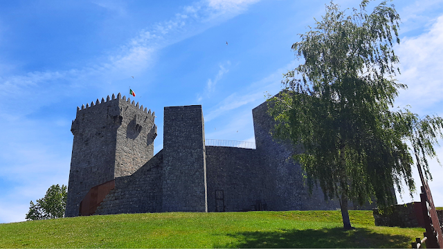 Vista exterior do Castelo de Montalegre, com muralhas em pedra, torre de menagem e árvore em primeiro plano, num dia de céu azul em Trás-os-Montes.