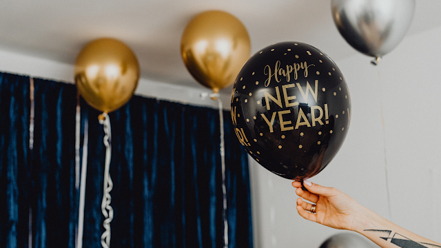 Celebração de Ano Novo com Balões Dourados Mão a segurar um balão preto com a mensagem Happy New Year, rodeado de balões dourados e prateados numa decoração festiva de Ano Novo.