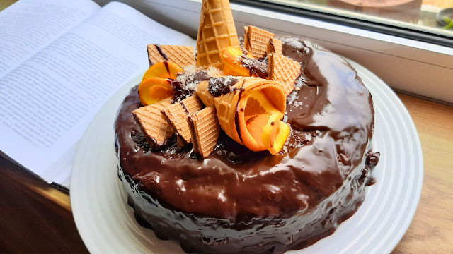 Bolo de laranja caseiro coberto com chocolate derretido, decorado com bolachas, rodelas de laranja e cone de gelado em cima.