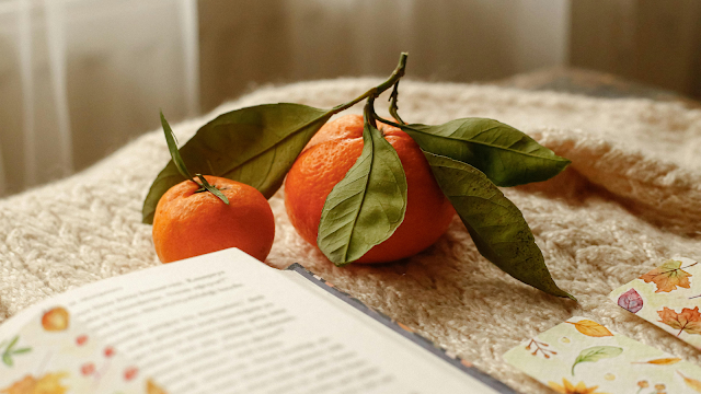 Fotografia de tangerinas frescas com folhas verdes sobre uma manta bege, ao lado de um livro aberto e marcadores com desenhos de folhas de outono, criando um ambiente acolhedor e outonal.