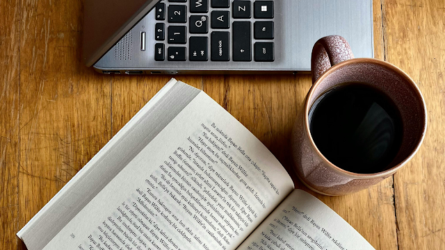 Livro aberto ao lado de um portátil e de uma chávena de café sobre uma mesa de madeira, simbolizando um momento de leitura e tranquilidade.