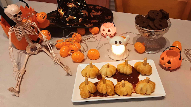 Mesa decorada para um jantar de Halloween, com mini pizas em formato de abóbora servidas num prato branco, decoração temática com esqueleto, abóboras luminosas, chapéu de bruxa, luzes decorativas e doces de chocolate.