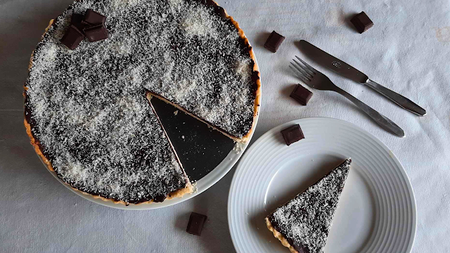 Tarte de chocolate com coco ralado – receita fácil e deliciosa Fatia de tarte de chocolate coberta com coco ralado servida num prato branco, acompanhada de quadrados de chocolate e talheres sobre uma toalha clara.