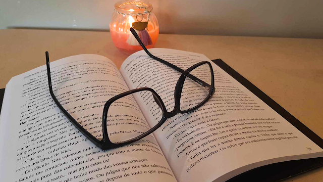 Óculos graduados sobre um livro aberto com uma vela acesa ao fundo, representando leitura, cansaço visual e rotina com astigmatismo