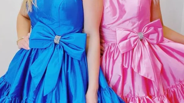 Detailed close-up showing two women standing side-by-side, wearing matching satin cocktail dresses with large statement bows at the waist, cinched by sparkling rhinestone rings. The woman on the left is in a vibrant royal blue dress, and the woman on the right is in a candy pink dress. Both dresses feature voluminous, tiered, and ruffled skirts. Only the dresses, bows, and a hint of the wearer's arms and shoulders are visible against a simple white studio background.