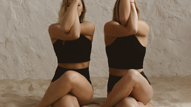 Imagem captada de frente de duas mulheres com roupas de desporto pretas, calções e tops, deitadas e de pernas cruzadas na areia. Ambas estão a realizar a pose de yoga da águia, com os braços entrelaçados em frente ao rosto e as mãos unidas. O fundo é uma parede texturizada de cor creme. A iluminação é suave e natural.
