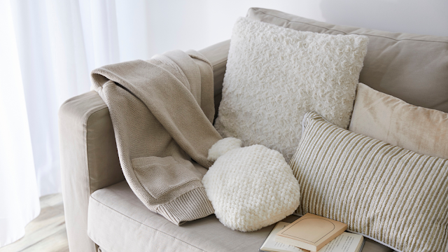 Sofá bege com almofadas macias, manta de malha e livro aberto, criando um ambiente acolhedor de soft living.