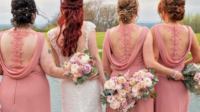 Foto de costas de uma noiva vestida de branco ao lado de três madrinhas vestindo vestidos longos em tom de rosa seco com detalhes em renda e transparência nas costas. Todas seguram buquês florais com tons de rosa, creme e verde, em um cenário externo com gramado e água ao fundo.