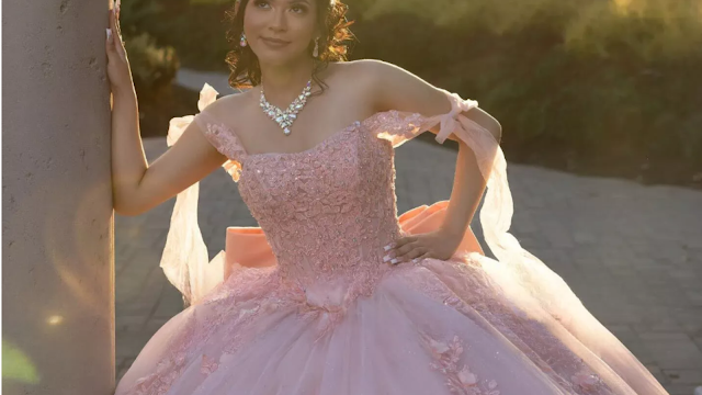 Uma jovem radiante com um deslumbrante vestido de Quinceañera rosa pastel. O corpete é rendado e brilhante, e as alças são fluidas e delicadas. Ela usa joias reluzentes e seu cabelo está penteado com carinho. Ela se apoia em uma coluna, olhando para o horizonte com um sorriso sonhador, enquanto a luz dourada do sol poente a envolve, criando uma atmosfera mágica e inesquecível.