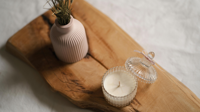 Vela aromática em frasco de vidro com tampa decorativa ao lado de um vaso cor-de-rosa com flores secas, sobre uma tábua de madeira rústica.