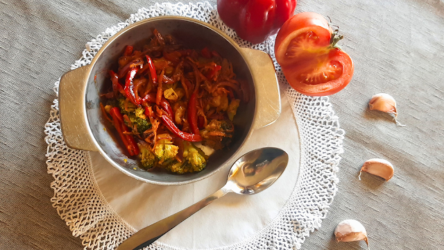 Prato colorido de legumes salteados servido numa tigela metálica sobre um naperon branco rendado. Os legumes incluem brócolos, tiras de pimento vermelho e cenoura. Ao lado, há uma colher de metal, um tomate cortado ao meio, um pimento vermelho inteiro e dentes de alho, tudo sobre uma toalha cinzenta.