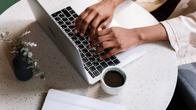 Mãos a escrever num computador portátil sobre uma mesa redonda, com uma chávena de café e uma pequena planta, num ambiente calmo e inspirador para criar conteúdos para um blog.