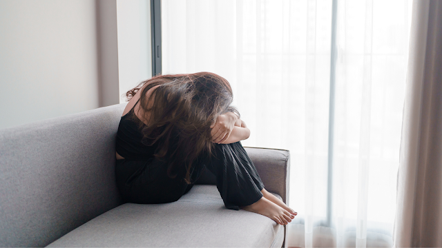 Mulher sentada num sofá com o corpo encolhido e a cabeça apoiada nos joelhos, transmitindo um momento de ansiedade e reflexão, junto a uma janela com luz suave.