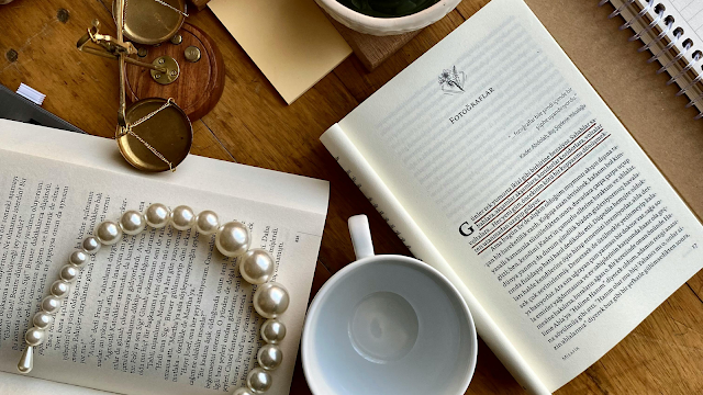 Livros abertos sobre uma mesa de madeira, acompanhados por uma chávena de café vazia, um colar de pérolas, um bloco de notas e óculos antigos, criando uma atmosfera de leitura e reflexão filosófica.