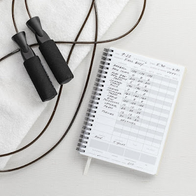 Planeamento de atividade física em casa Corda de saltar e caderno de registo de treino com exercícios anotados, representando o planeamento e a criação de uma rotina de atividade física.
