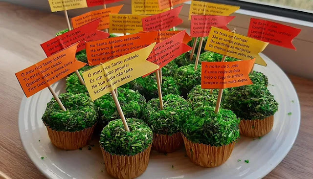 Uma travessa branca com vários brigadeiros cobertos de coco ralado tingido de verde vibrante, assemelhando-se a manjericos. Cada brigadeiro está numa forminha de papel castanha e decorado com uma pequena bandeirola de papel laranja ou amarela espetada num palito, contendo quadras populares portuguesas impressas. O fundo é luminoso e desfocado.