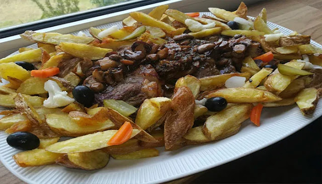 Uma travessa branca retangular servida com fatias de bife do vazio grelhado, cobertas com cogumelos salteados e um molho escuro, rodeadas por batatas fritas em palitos largos. A guarnição inclui azeitonas pretas inteiras, pedaços de couve-flor branca e fatias de picles e cenoura. A travessa está sobre uma mesa de madeira, perto de uma janela.