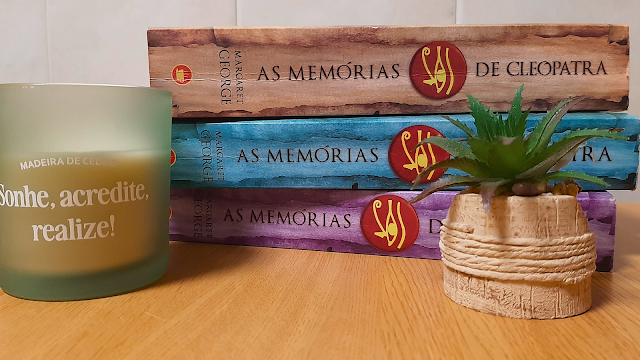 A Trilogia de Cleópatra e Inspiração Uma fotografia de plano médio sobre uma mesa de madeira clara. Empilhados horizontalmente, encontram-se os três volumes da trilogia 'As Memórias de Cleópatra' de Margaret George, com lombadas em tons de castanho, azul e roxo. À esquerda, uma vela de vidro fosco verde com a frase 'Sonhe, acredite, realize!'. À direita, uma pequena planta suculenta num vaso rústico que imita um barril com cordas.
