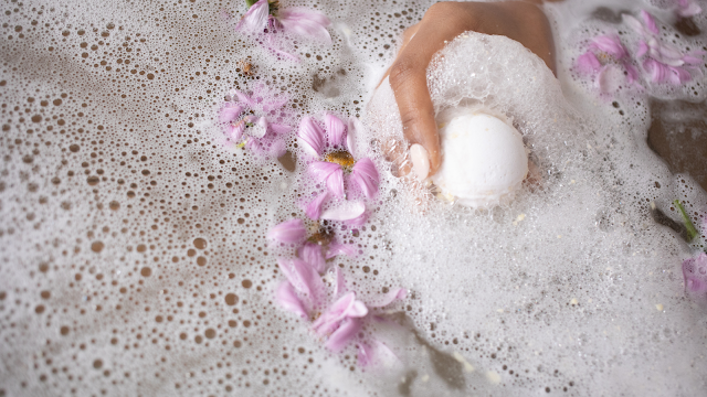 Mão segurando uma bomba efervescente num banho de espuma com pétalas de flores cor-de-rosa, criando um ambiente relaxante e de autocuidado.