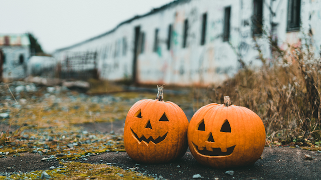 Duas abóboras de Halloween esculpidas com caras sorridentes, colocadas lado a lado num espaço exterior abandonado, criando um ambiente misterioso.