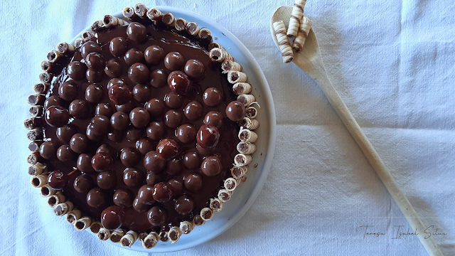 Bolo de chocolate, visto de cima sobre um fundo branco. o Bolo tem no seu topo bolas de chocolate e à sua volta biscoitos. Ao lado do bolo uma colher de pau com biscoitos decora a mesa.