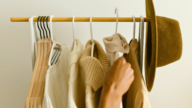Mão a escolher roupa em tons neutros num cabide com camisolas e chapéu castanho, representando estilo e organização no guarda-roupa de outono