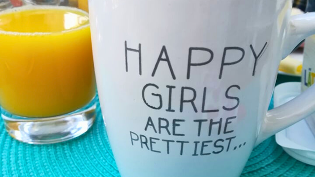 Caneca branca com a frase 'Happy girls are the prettiest' ao lado de um copo de sumo de laranja sobre uma toalha azul. A imagem transmite um momento de tranquilidade e bem-estar, alinhado com a prática de mindfulness e presença no momento.