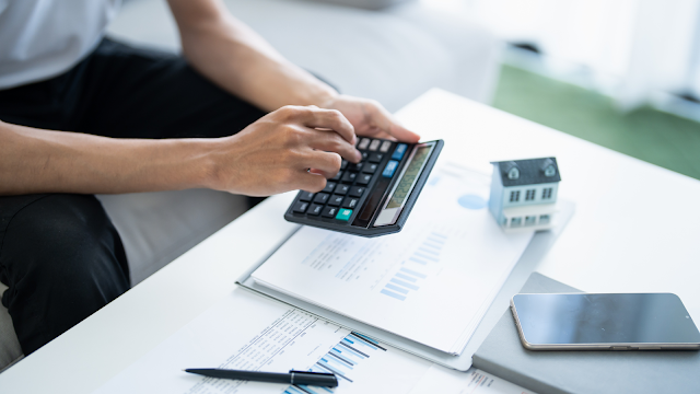 Pessoa a calcular despesas com habitação usando uma calculadora, com documentos financeiros e uma pequena casa em miniatura sobre a mesa.