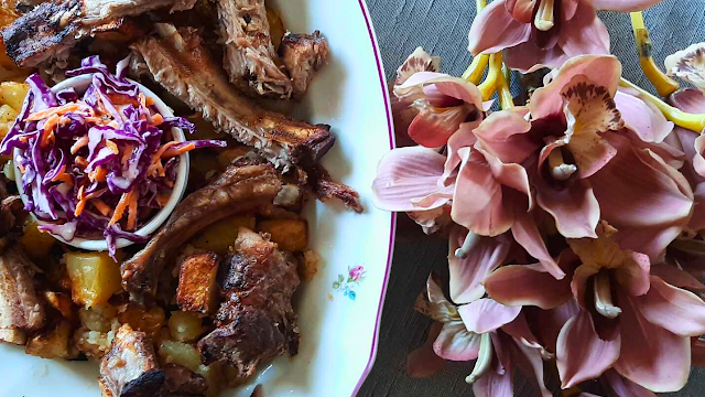 Costelinhas de porco assadas no forno com batatas douradas e salada de couve roxa, servidas numa travessa, ao lado de flores decorativas