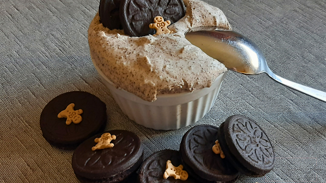 Pequena tigela com mousse de oreo no sei interior, em cima uma bolacha de oro a decorar. Na mesa, várias bolaschas oreia complementam a decoração juntamente com uma colher de sobremesa.