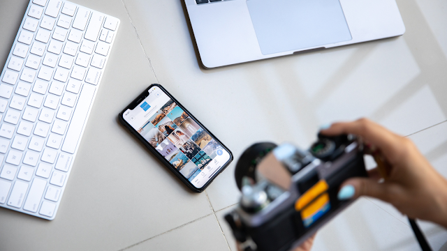 Teclado branco. telemóvel, com a tela a mostrar as publicações do Instagram, e uma mão a segurar uma máquina fotográfica