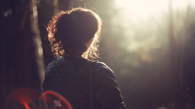 Mulher de costas, com cabelo castanho preso, vestindo casaco escuro, a olhar para a luz do sol filtrada entre árvores, numa atmosfera calma e introspectiva ao ar livre.