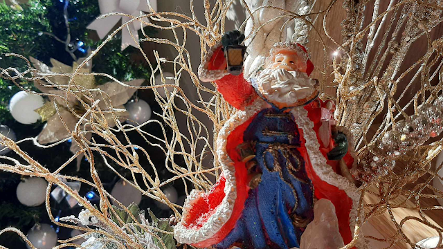 O Brilho da Inocência: À Procura do Pai Natal Figura decorativa de um Pai Natal clássico com fato vermelho e azul, segurando uma lanterna acesa. Ele está rodeado por ramos decorativos dourados e cintilantes, com uma árvore de Natal desfocada ao fundo, decorada com bolas brancas.