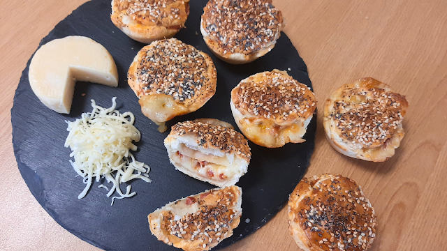 Empadas de queijo e chouriço douradas, cobertas com sementes de sésamo e papoila, servidas num prato de ardósia ao lado de queijo e queijo ralado.