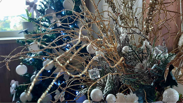 Grande plano focado num arranjo decorativo de Natal composto por ramos finos e ondulados cobertos de purpurina dourada. No centro, destacam-se pequenas esferas brancas com textura de neve e cubos de acrílico transparentes. Ao fundo, de forma desfocada, vislumbra-se uma árvore de Natal verde decorada com luzes azuis e bolas brancas, criando um ambiente festivo e sofisticado.