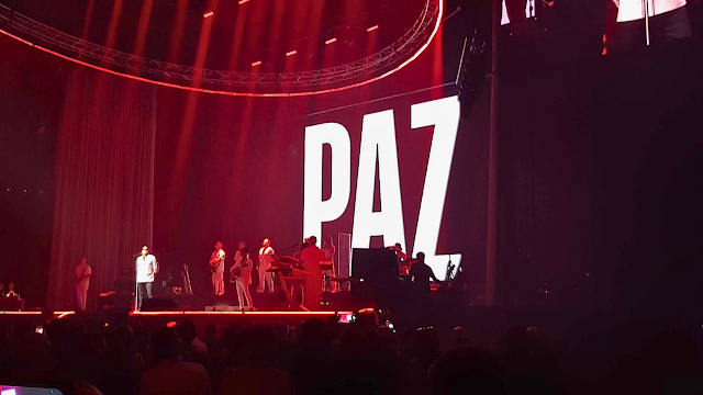 Fotografia de palco do concerto de Pedro Abrunhosa na Super Bock Arena. O artista, vestido de branco, está no centro com a sua banda. O palco está iluminado com luzes vermelhas intensas e, num grande ecrã de fundo, a palavra "PAZ" aparece escrita em letras brancas maiúsculas e grandes. Em primeiro plano, vê-se a silhueta do público a assistir ao espetáculo.
